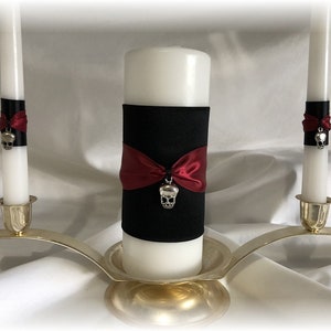 May include: Three white candles with black and burgundy ribbon accents and silver skull charms. The candles are on a gold metal stand.
