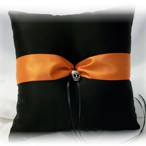 May include: Black satin pillow with an orange satin ribbon and a silver skull accent. The pillow is designed for wedding rings.
