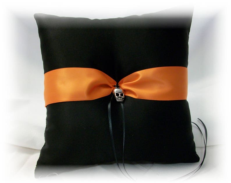 May include: Black satin pillow with an orange satin ribbon and a silver skull accent. The pillow is designed for wedding rings.