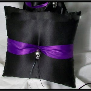 May include: Black satin pillow with a purple ribbon and a silver skull charm. The pillow is designed for a wedding ceremony.