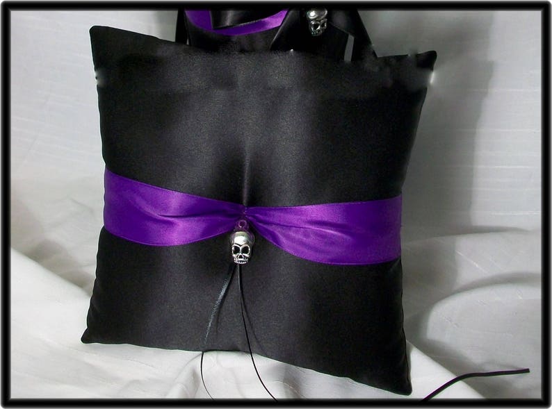 May include: Black satin pillow with a purple ribbon and a silver skull charm. The pillow is designed for a wedding ceremony.