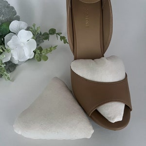 Puede incluir: Un par de sandalias de tacón de cuero marrón con puntera abierta y correa de tobillo. Las sandalias están sobre una superficie blanca con un triángulo de tela blanca y un cojín de tela blanca en forma de corazón.