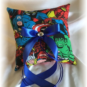 May include: A blue and white ring pillow with a comic book design featuring superheroes like Captain America, Hulk, and Spiderman. The pillow has a blue satin ribbon tied in a bow with a rhinestone in the center.