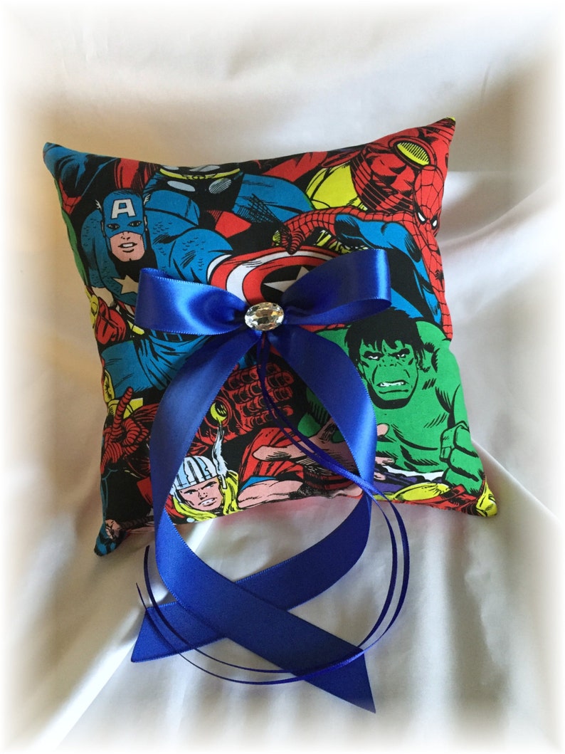 May include: A blue and white ring pillow with a comic book design featuring superheroes like Captain America, Hulk, and Spiderman. The pillow has a blue satin ribbon tied in a bow with a rhinestone in the center.