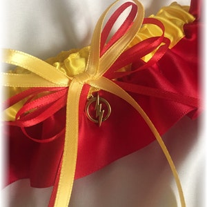 May include: A red and yellow garter with a gold lightning bolt charm. The garter features a yellow bow and red and yellow ribbons. The garter is made of satin-like material and is likely for a wedding or special occasion.