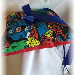 May include: A heart-shaped pillow with a red border and a blue ribbon. The pillow is covered in a colorful fabric featuring various superhero characters, including Captain America, Iron Man, and Spider-Man.