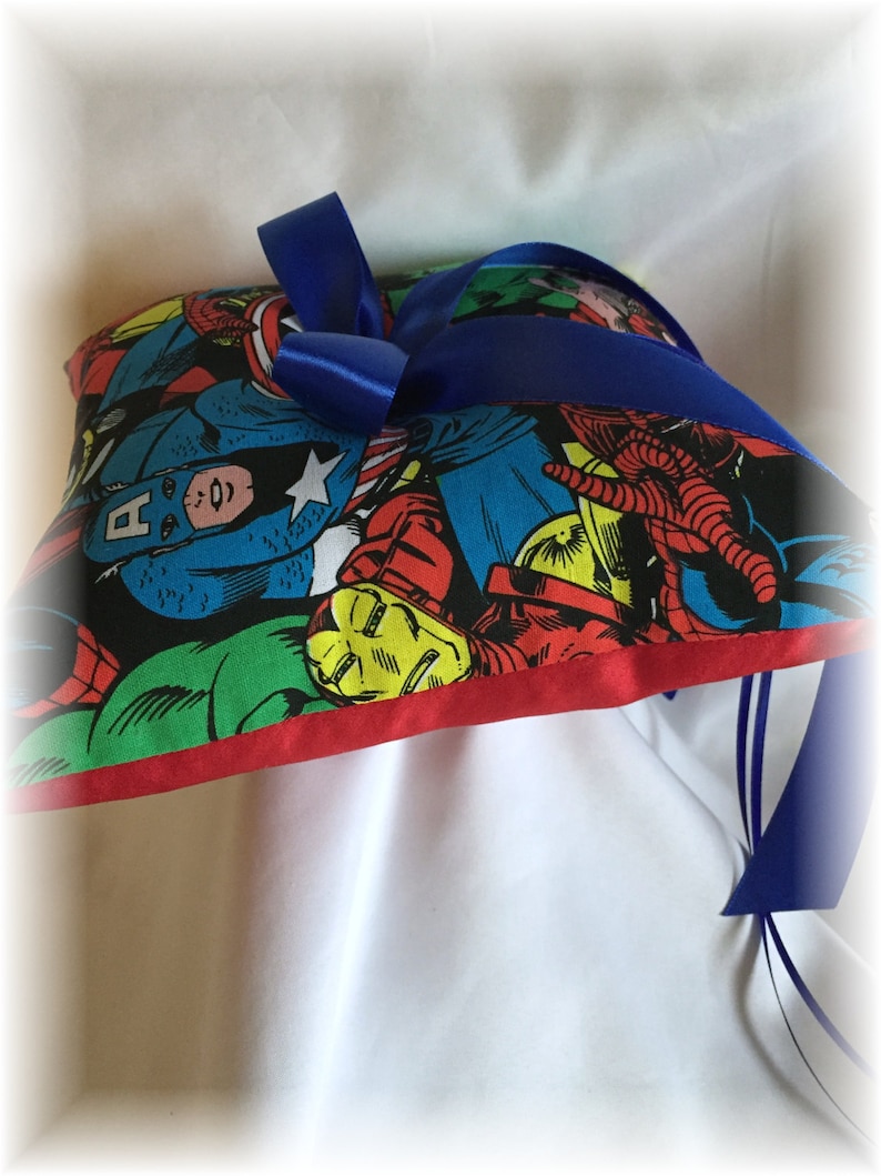 May include: A heart-shaped pillow with a red border and a blue ribbon. The pillow is covered in a colorful fabric featuring various superhero characters, including Captain America, Iron Man, and Spider-Man.