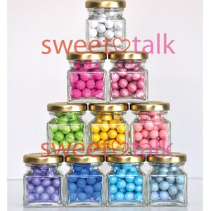 Pode incluir: Uma pilha de dez pequenos frascos de vidro com tampas douradas cheios de doces coloridos. Os frascos est&atilde;o dispostos em forma de pir&acirc;mide. Os doces s&atilde;o de v&aacute;rias cores, incluindo branco, rosa, verde, amarelo, roxo, azul e azul claro.