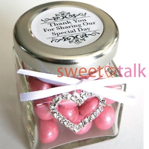 Pode incluir: Um pequeno frasco de vidro cheio de doces cor-de-rosa, amarrado com uma fita branca e um amuleto de cora&ccedil;&atilde;o prateado. O frasco tem uma tampa prateada com um r&oacute;tulo branco que diz "Obrigado por partilhar o nosso dia especial".