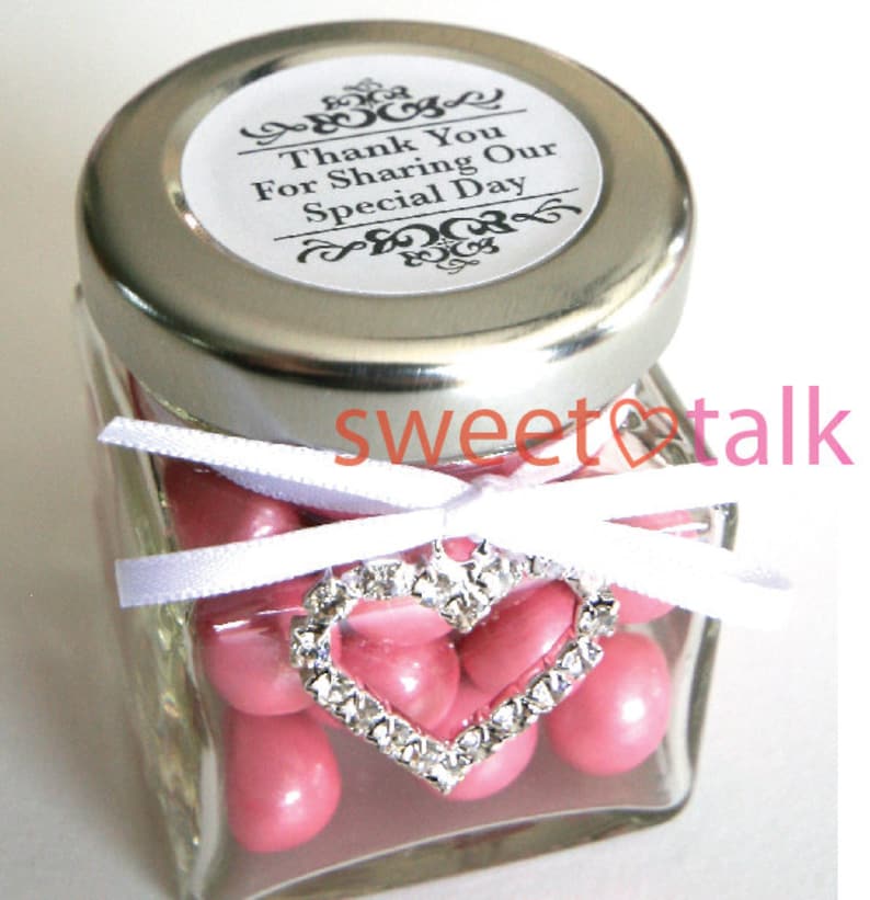 Pode incluir: Um pequeno frasco de vidro cheio de doces cor-de-rosa, amarrado com uma fita branca e um amuleto de cora&ccedil;&atilde;o prateado. O frasco tem uma tampa prateada com um r&oacute;tulo branco que diz "Obrigado por partilhar o nosso dia especial".