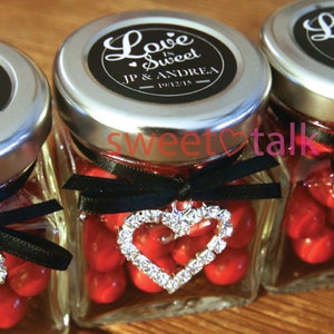 Pode incluir: Pequenos frascos de vidro cheios de doces vermelhos, amarrados com uma fita preta e um charme de cora&ccedil;&atilde;o prateado. As tampas dos frascos s&atilde;o prateadas com etiquetas pretas e brancas que dizem "Love is Sweet" com os nomes JP & Andrea e a data 10/12/15.