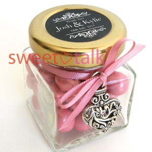 Pode incluir: Um pequeno frasco de vidro cheio de doces cor-de-rosa, amarrado com uma fita rosa e um amuleto de cora&ccedil;&atilde;o prateado. A tampa &eacute; dourada com um r&oacute;tulo preto que diz "Obrigado Josh & Kylie 18 de julho de 2015".