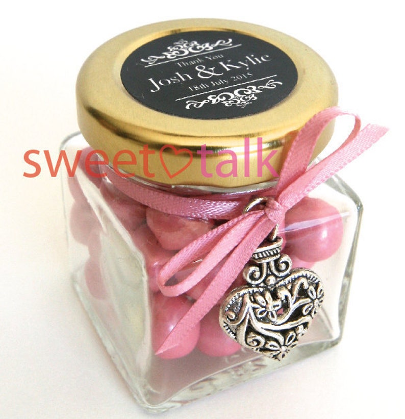 Pode incluir: Um pequeno frasco de vidro cheio de doces cor-de-rosa, amarrado com uma fita rosa e um amuleto de cora&ccedil;&atilde;o prateado. A tampa &eacute; dourada com um r&oacute;tulo preto que diz "Obrigado Josh & Kylie 18 de julho de 2015".