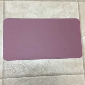 May include: A rectangular, mauve-colored, non-slip mat. The mat is made of a textured material.