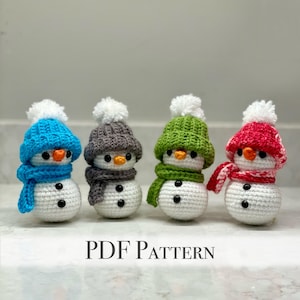 以下が含まれることがあります： 異なる色の帽子とマフラーをつけた、手編みの雪だるまのオーナメントが4つ。雪だるまは白で、目は黒く、ボタンがついています。帽子はグレー、グリーン、ブルー、ピンク。マフラーはブルー、グリーン、グレー、レッドです。