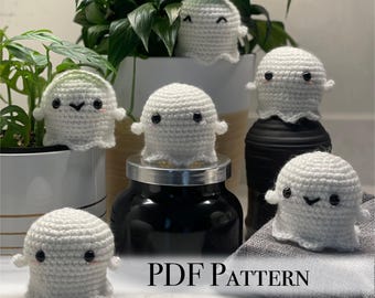 May include: A set of six crocheted ghost amigurumi toys with black eyes and smiling faces. The ghosts are sitting on a variety of surfaces, including a plant pot, a jar, and a towel. The image includes the text "PDF PATTERN".