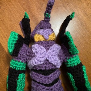 May include: Crocheted plush toy of a purple and green robot with a yellow face. The robot has a large, purple butterfly-shaped chest piece.