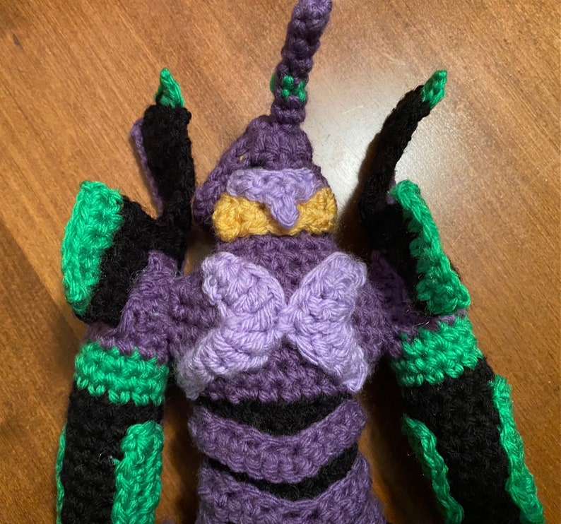 May include: Crocheted plush toy of a purple and green robot with a yellow face. The robot has a large, purple butterfly-shaped chest piece.