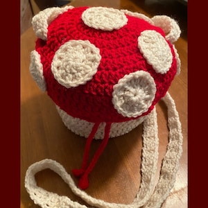 May include: A crocheted mushroom-shaped bag with a white strap. The bag is red with white circles on the top.