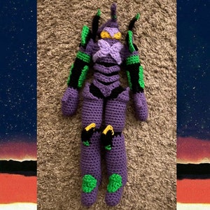 May include: A crocheted plush toy of a purple robot with green and black accents. The robot has a large head with a yellow eye and a purple and black body.
