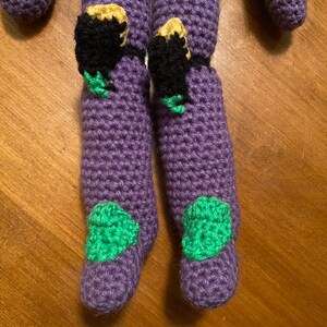 May include: Purple crocheted legs with green and black details. The legs are made of yarn and have a textured surface.