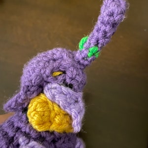 May include: Purple crocheted stuffed toy with a yellow beak and green accents on the head.