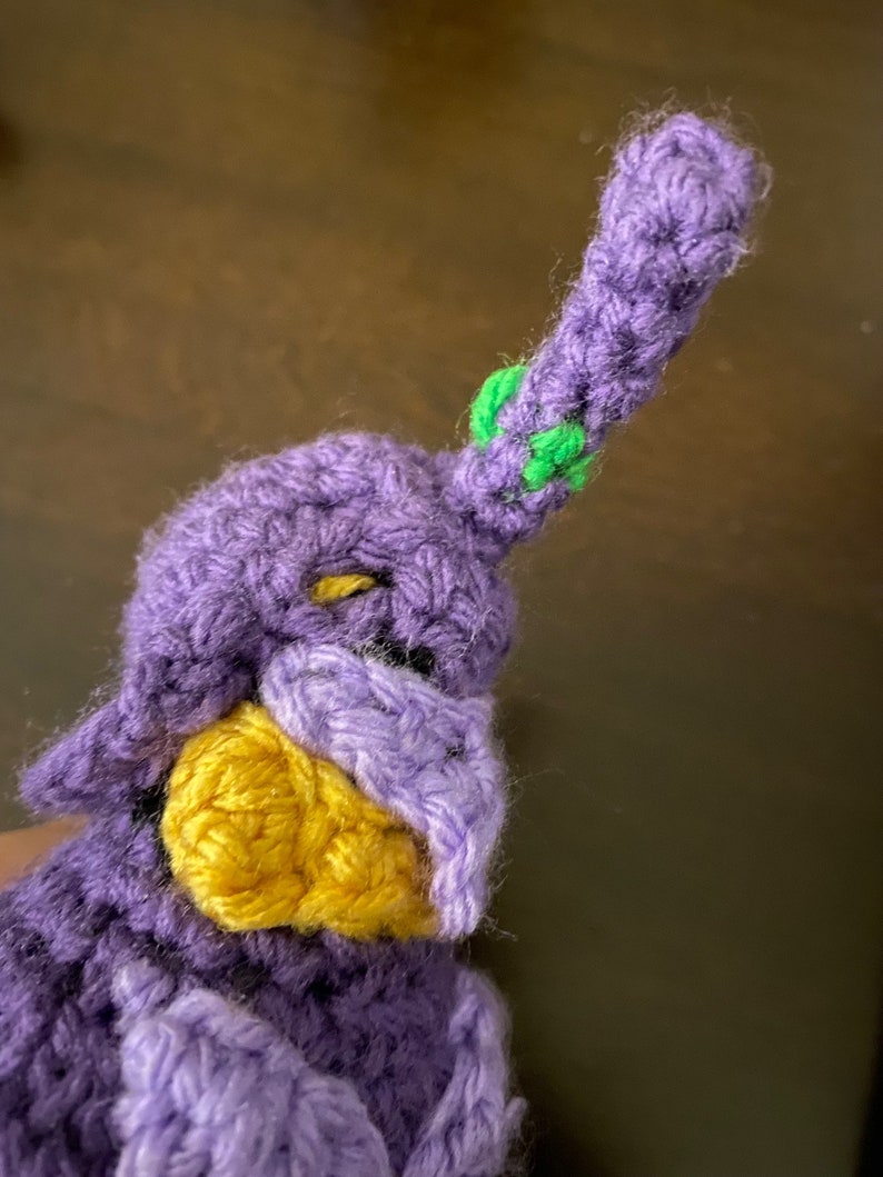 May include: Purple crocheted stuffed toy with a yellow beak and green accents on the head.