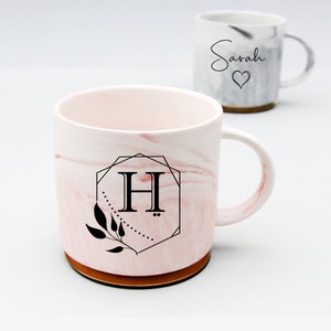 Op de afbeelding: Een roze en witte marmeren mok met een gouden rand en een zwart geometrisch ontwerp met de letter "H" en een bladmotief. De mok heeft een bijpassende schotel.