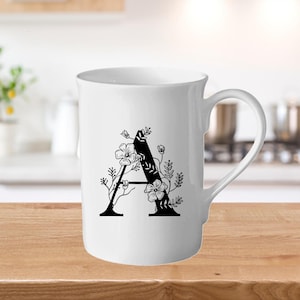 Könnte beinhalten: Weiße Keramiktasse mit einem schwarzen floralen "A"-Design. Die Tasse hat einen gebogenen Griff und eine einfache Form. Das florale Design umfasst Blumen und Blätter.