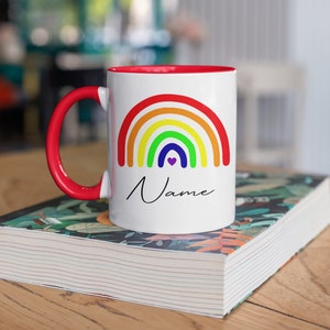 Op de afbeelding: Witte keramische mok met een rode rand en handvat. De mok heeft een regenboogontwerp met een klein hartje in het midden en het woord "Naam" eronder geschreven. De mok staat op een boek met een bloemenpatroon.