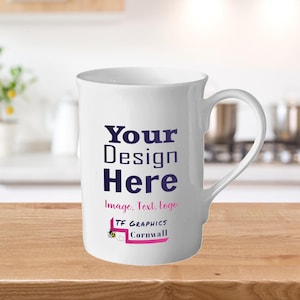 Può includere: Tazza in ceramica bianca con manico curvo. La tazza presenta il testo "Your Design Here" in blu scuro, con "Image. Text. Logo" sotto. Il fondo della tazza riporta la scritta "TF Graphics Cornwall" in rosa e nero.