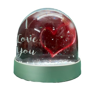 Op de afbeelding: Een transparante glazen sneeuwbol met een rood hart en de woorden "I Love You" in witte letters. De basis van de sneeuwbol is lichtgroen.