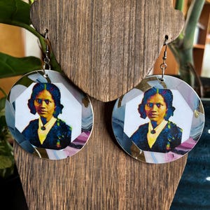 May include: A pair of round dangle earrings featuring a central portrait of a person with dark hair wearing a dark jacket and white collared shirt. The portrait is framed by a circular border with a marbled pattern in shades of blue, purple, and white.
