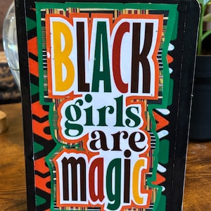 Puede incluir: Una libreta negra con un diseño colorido que incluye el texto "Black girls are magic". El diseño incluye un fondo estampado en verde, naranja y amarillo.