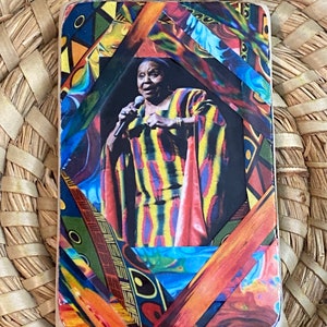 Peut inclure: Un dessous de verre en bois coloré avec un portrait d'une femme tenant un microphone. La femme porte une robe rayée jaune, rouge et bleue. Le dessous de verre a un motif géométrique avec des couleurs orange, jaune, bleu et vert.