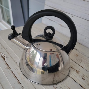 May include: A silver kettle with a black handle and a black knob on the lid. The kettle is sitting on a white wooden surface.