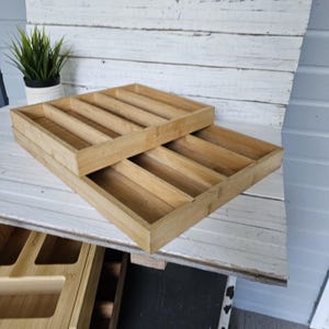 May include: Two rectangular bamboo utensil organizers with multiple compartments. The organizers are stacked on a white wooden surface, with a small potted plant in the background. The organizers are made of light-colored bamboo.