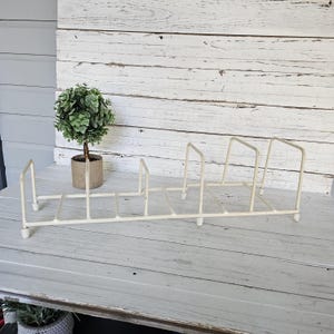 May include: Off-white metal plate rack with a rectangular base and vertical dividers. The rack is designed to hold plates upright. A small potted plant sits to the left of the rack on a white wooden surface.
