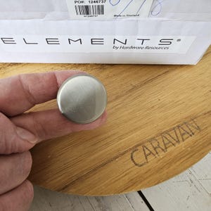 May include: A brushed nickel cabinet knob held in a hand, with a round, flat design. The knob is set against a wooden surface with the word "CARAVAN" etched into it. The packaging label reads "ELEMENTS by Hardware Resources".