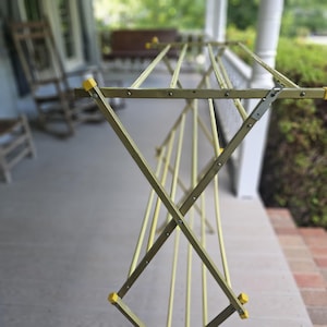 May include: An open, vintage-style clothes drying rack. The rack is made of metal with a pale yellow finish and yellow plastic end caps. The rack is set up on a porch, with a blurred background of a house and trees.