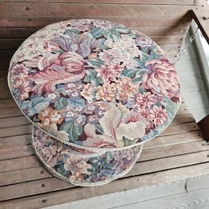 May include: A two-tiered, round stool with a floral tapestry design. The top and base feature a repeating pattern of pink, purple, blue, and beige flowers on a dark background. The edges are trimmed with a cream-colored, textured border. The stool sits on a wooden surface.
