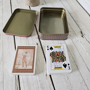 May include: A vintage playing card set with a golf-themed card and a King of Spades card, next to an open rectangular tin. The tin has a woven pattern on the sides and a gold interior. The lid is off to the side.