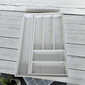 May include: A white wooden cutlery tray with multiple compartments. The tray has a long, narrow section on the left and several smaller, divided sections on the right. The tray is designed to organize kitchen utensils and is made of painted wood.