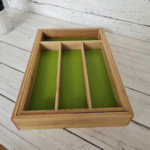 May include: A bamboo cutlery tray with a bright green felt interior. The tray is divided into four compartments and has the words "cook's tools" etched on the front. The tray sits on a white wooden surface.