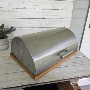 May include: A stainless steel bread box with a semi-cylindrical roll-top lid and a horizontal metal handle. The container is mounted on a rectangular wooden base with a light brown finish, featuring a smooth, metallic surface and clean lines.