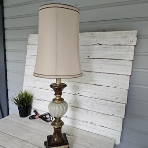 May include: A table lamp featuring a tall, beige fabric shade with dark trim. The base consists of a dark brown, textured square pedestal, a central white glass orb with a vertical ribbed pattern, and metallic gold accents connecting the parts.