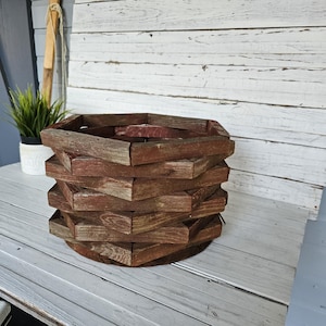 May include: A rustic, multi-layered wooden basket with a hexagonal design. The basket is constructed from weathered, reddish-brown planks, giving it a vintage appearance. The basket sits on a round, dark brown base, and is suitable for home decor.