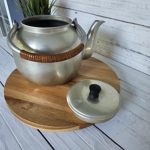 May include: A vintage silver-colored metal teapot with a wooden handle and a matching lid with a black knob. The teapot is sitting on a round wooden surface.
