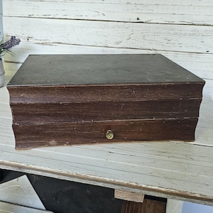 May include: A rectangular dark brown wooden storage box with a flat top and a single front drawer. The drawer features a small, centered circular brass knob. The box has a slightly curved, decorative molding along the front edge of the lid and base.