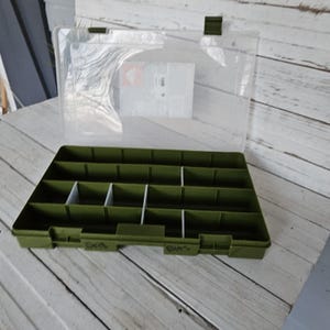 May include: An open, olive-green plastic storage box with a clear lid. The box is divided into multiple compartments by white dividers. The lid is attached with a dark green hinge and clasp. The words "BITS" and "DRILLS" are written on the box.